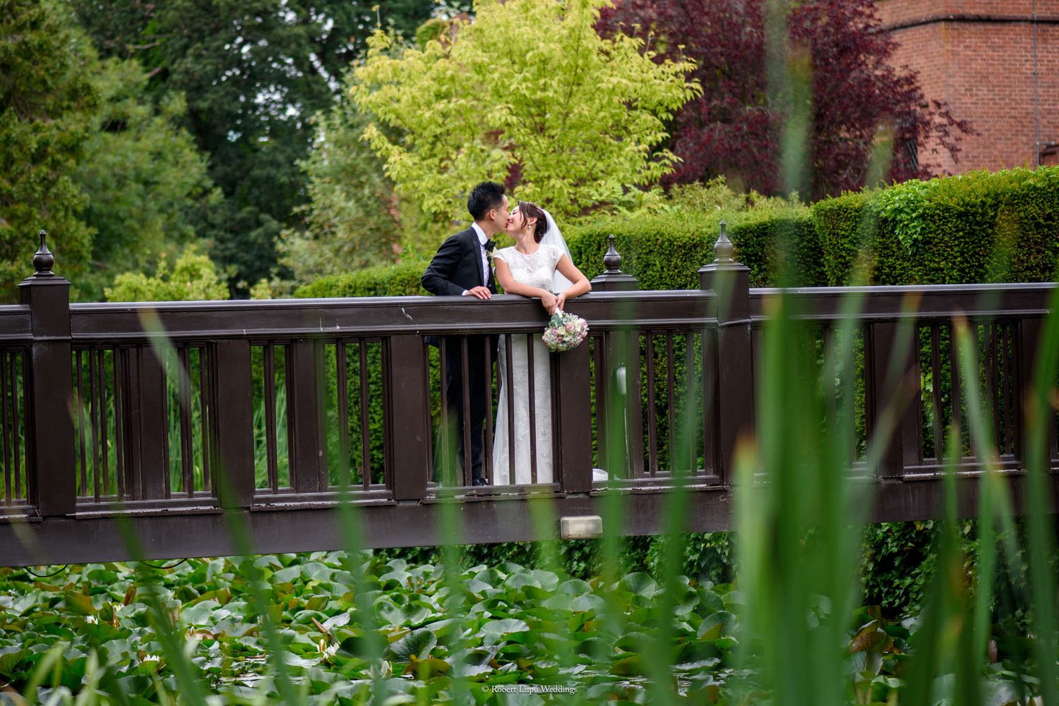 A bride and groom share a romantic moment on a wooden bridge surrounded by lush green gardens and a tranquil pond.