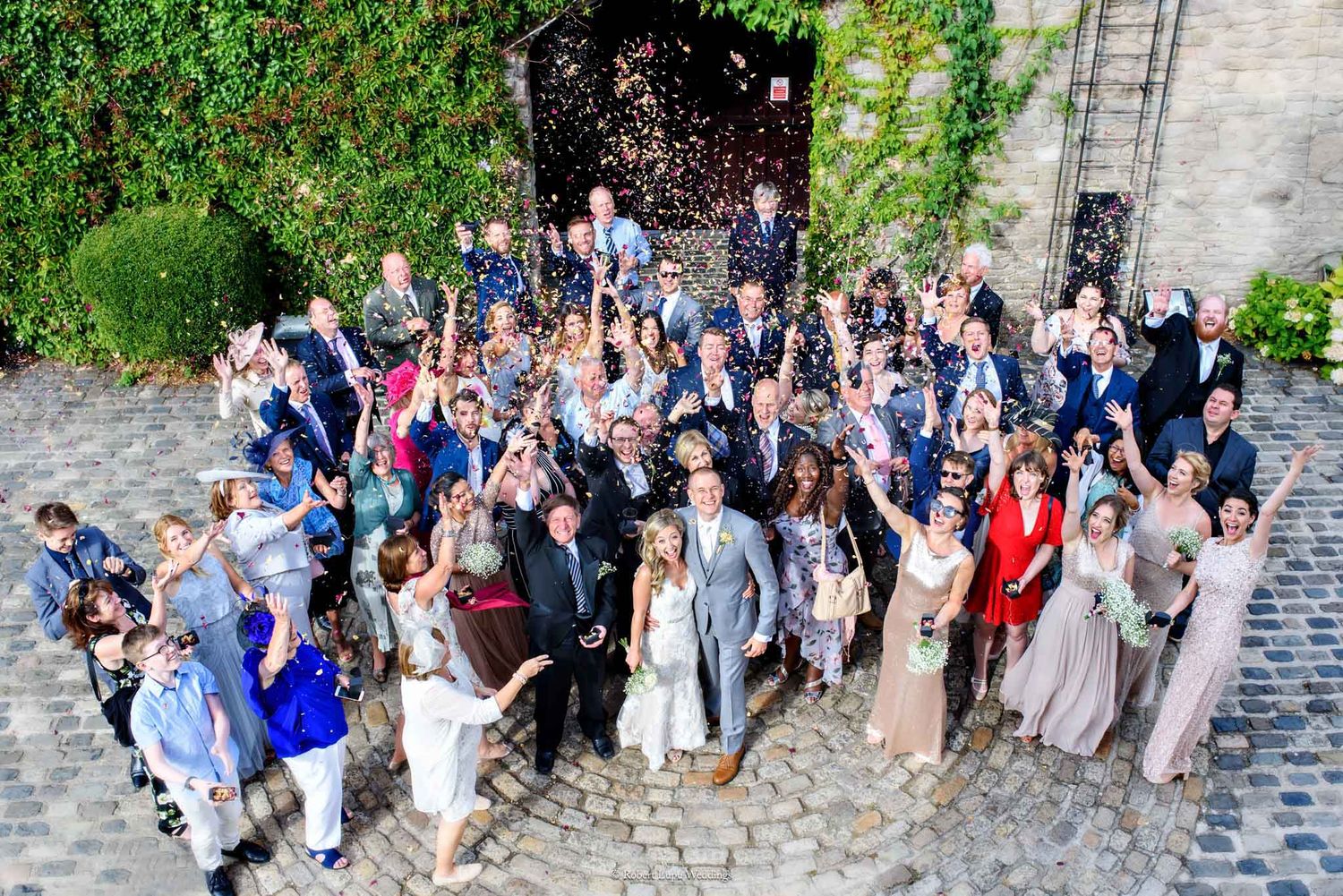 Large wedding group photo with confetti celebration outside a historic stone building on a sunny day.