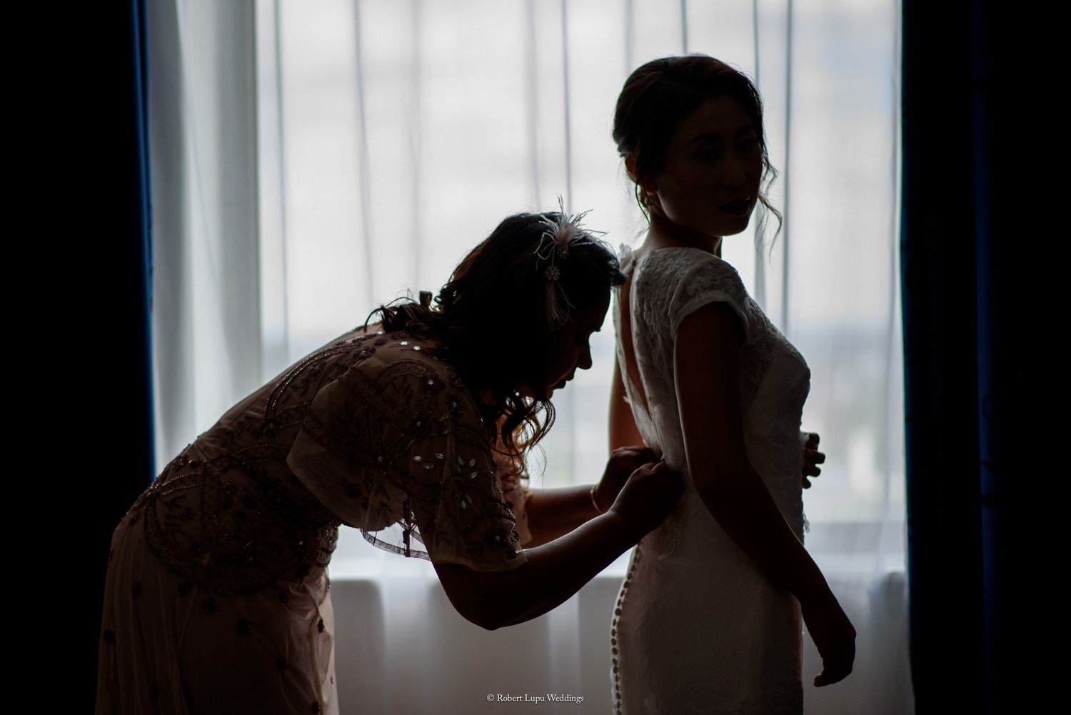 Silhouette of a bridesmaid helping the bride with her wedding dress in front of a bright window.