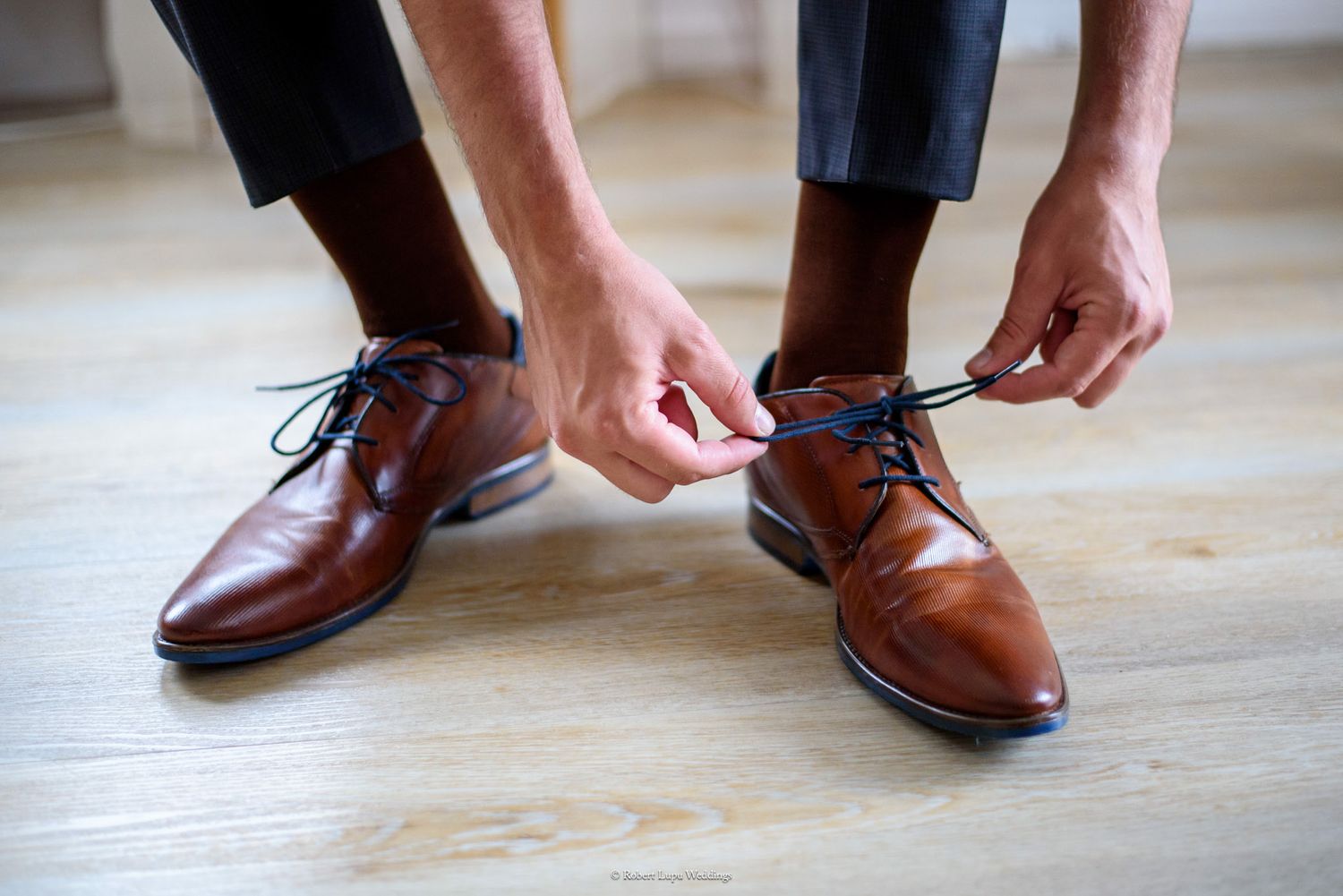 Man tying brown leather dress shoes with dark laces on a light hardwood floor.