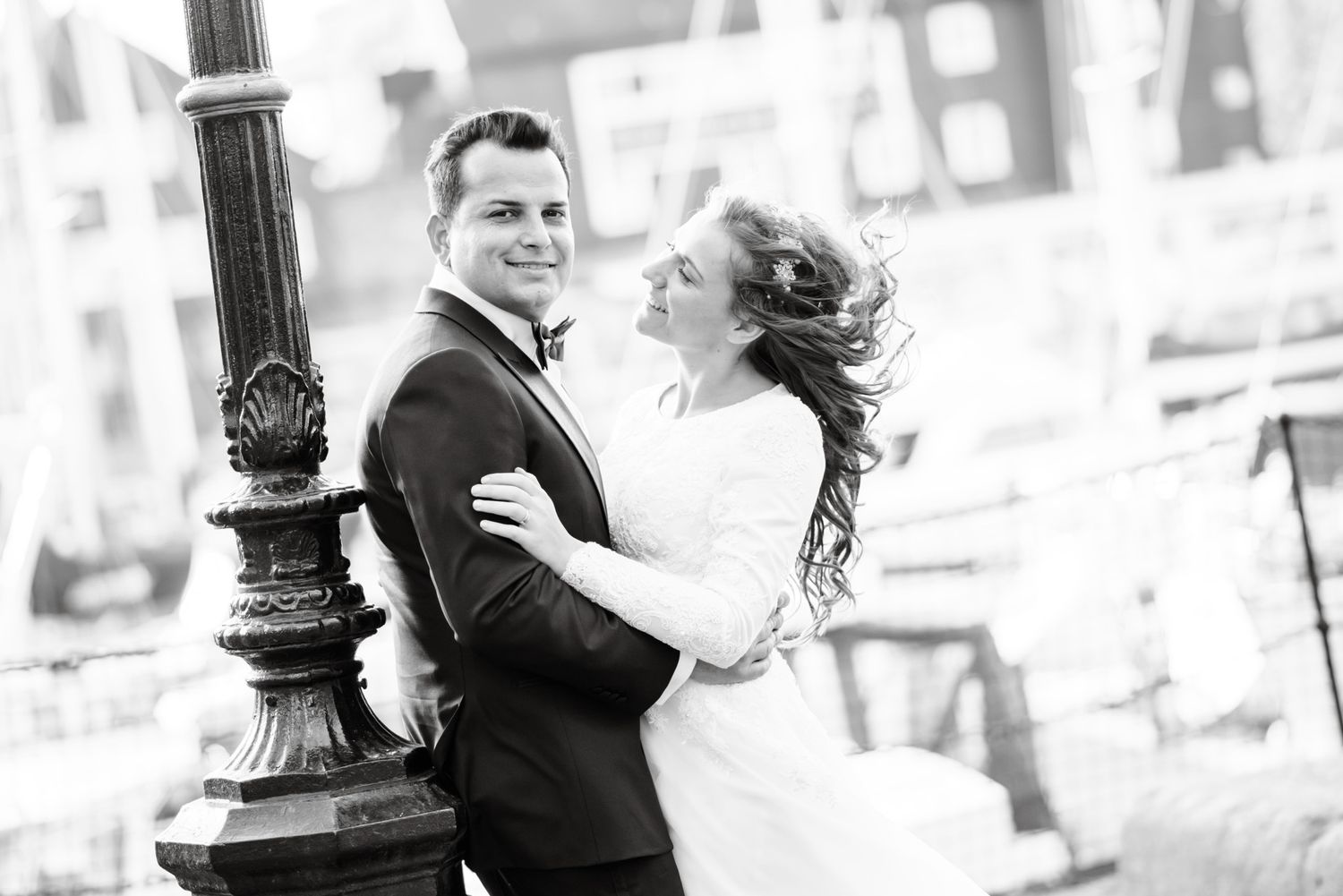 Black and white wedding photo of a smiling couple embracing near an ornate street lamp outdoors.