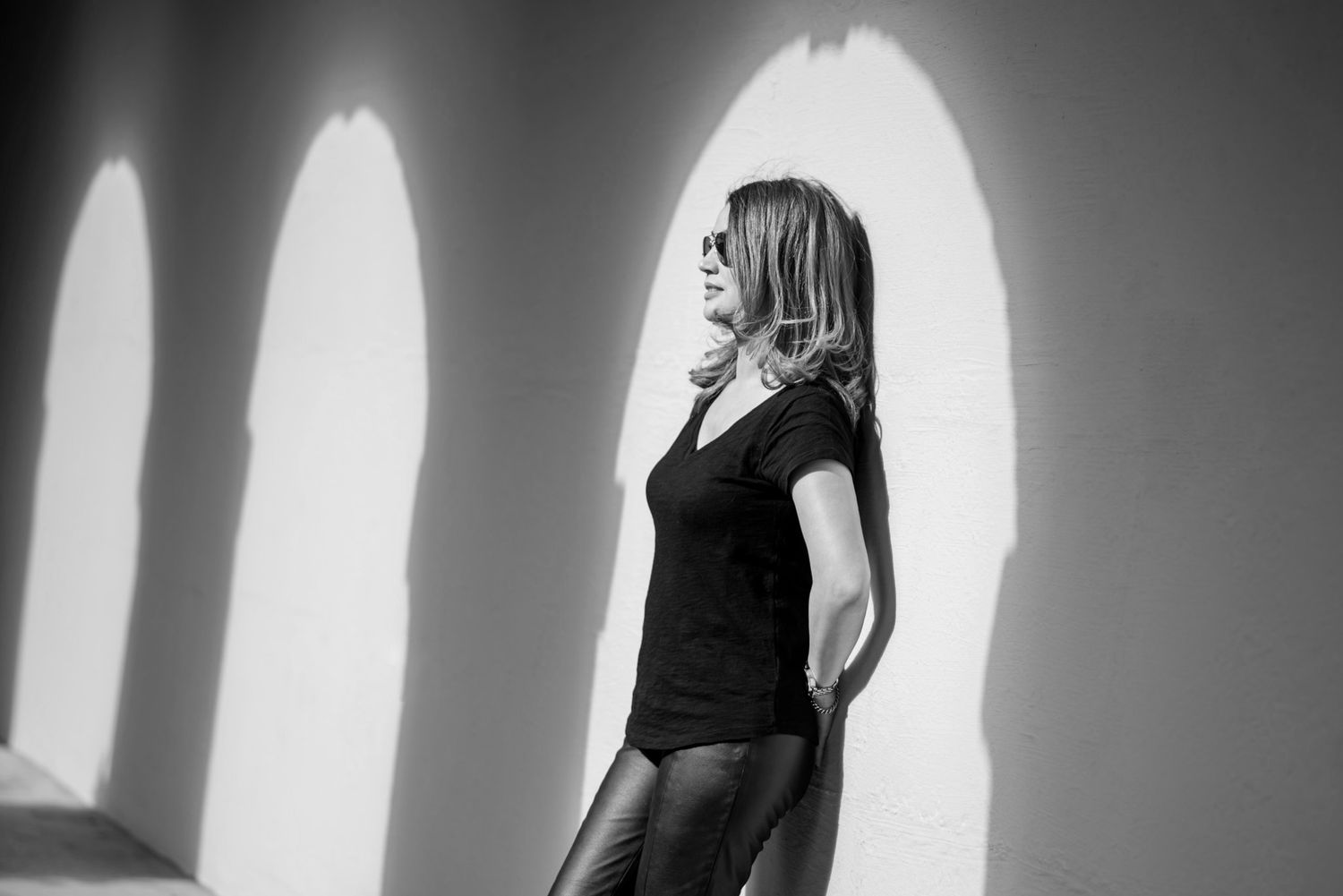 Black and white photo of a woman in a black top and leather pants posing against a wall with arched shadows.