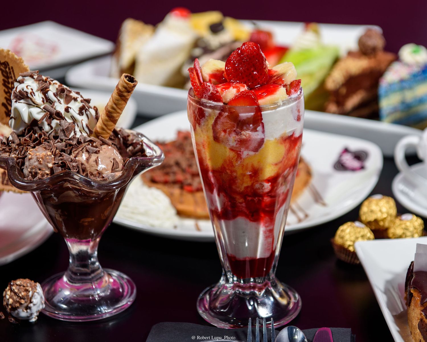 Decadent ice cream sundaes with chocolate and strawberry toppings served in tall glasses on a dessert table.
