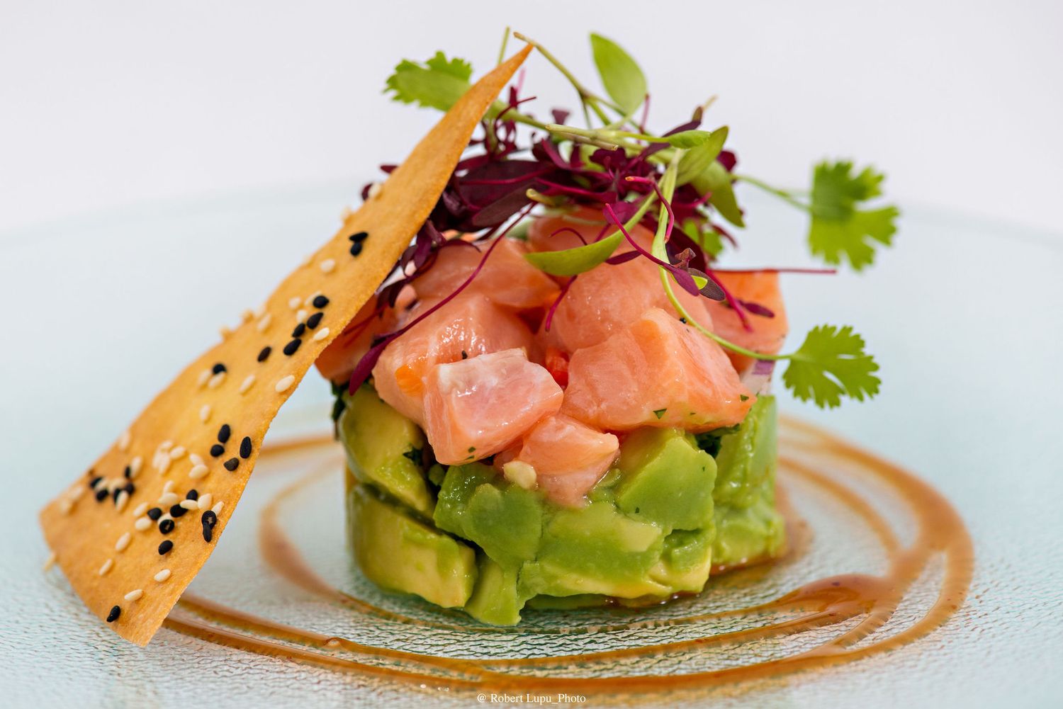 Elegant salmon and avocado tartare topped with microgreens and sesame crisp on a white plate.
