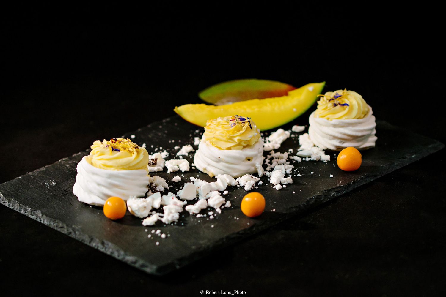 Elegant meringue desserts with mango curd on slate board, garnished with orange pearls and fresh mango slices.