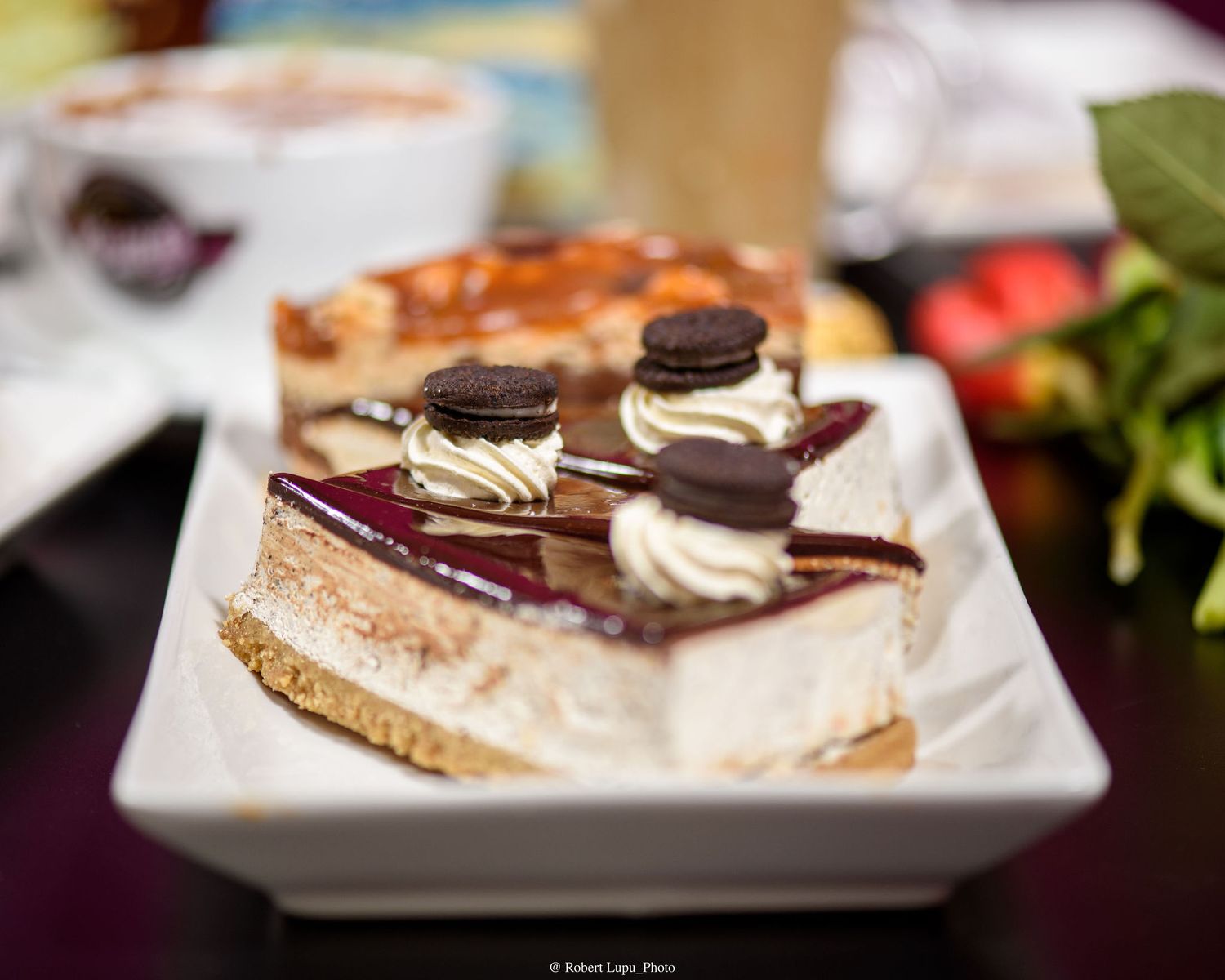 A slice of cheesecake topped with whipped cream, Oreo cookies, and berry glaze on a white square plate.