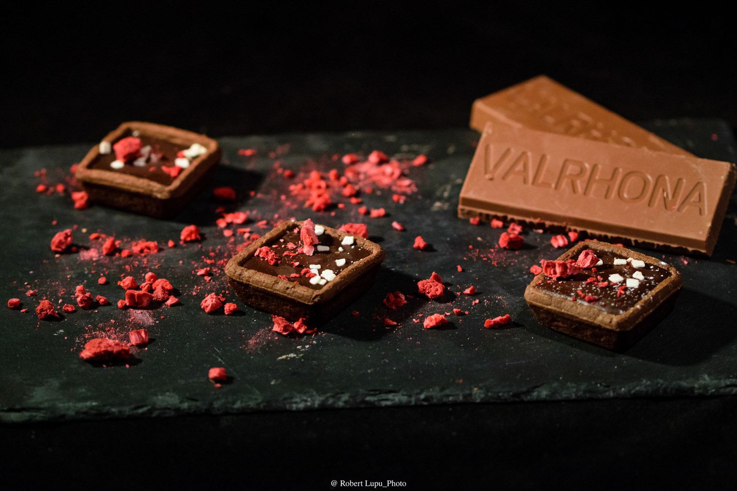 Valrhona chocolate bar pieces topped with freeze-dried raspberries displayed on a dark slate surface.