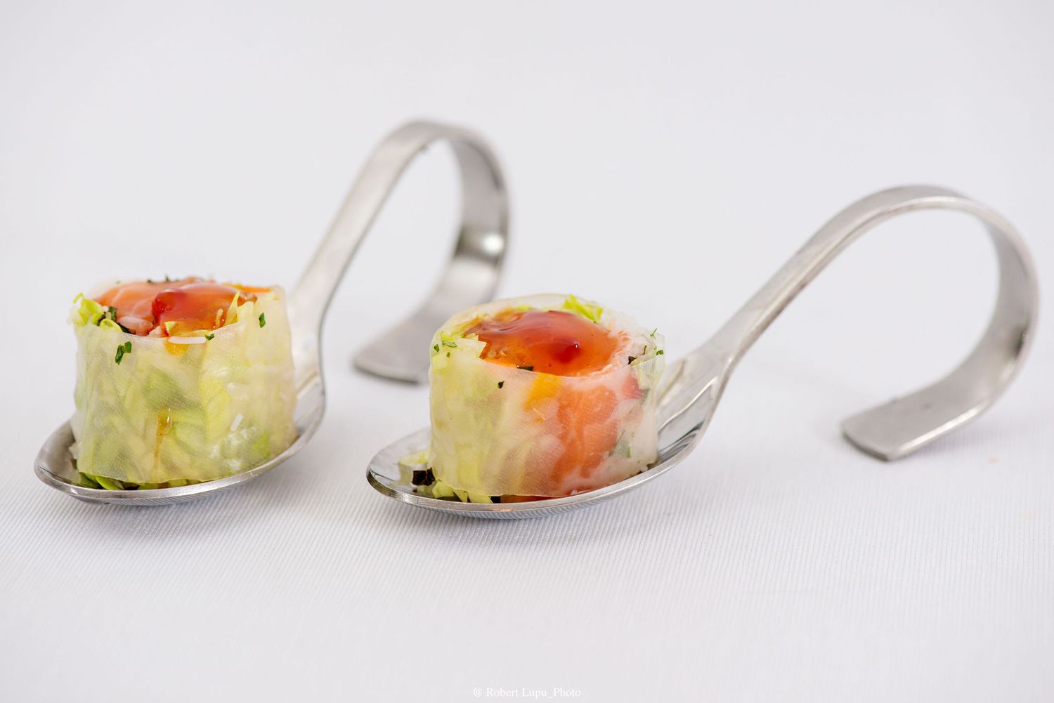 Two silver tasting spoons with elegant smoked salmon and avocado amuse-bouche appetizers on white background.