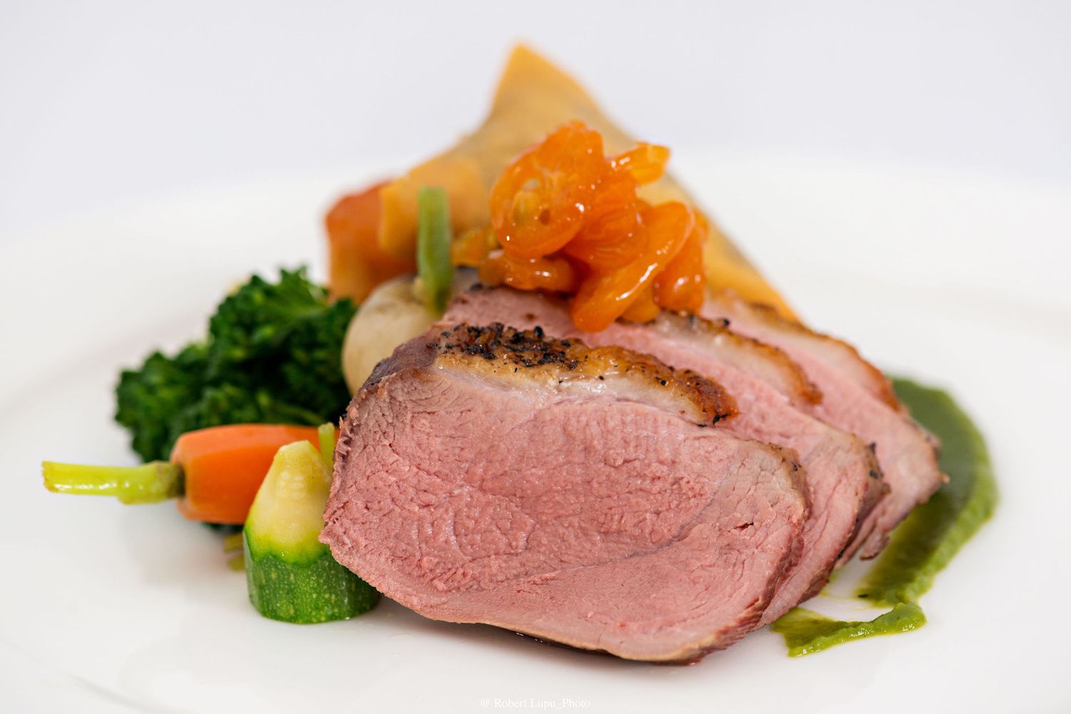 Sliced roasted lamb with kale, glazed carrots, zucchini, and apricot chutney on a white plate.