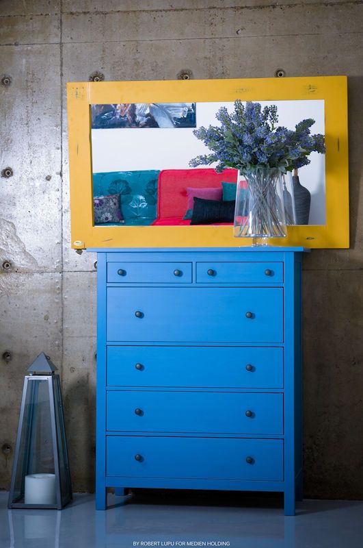 Bright blue dresser with yellow-framed mirror and purple flowers in a modern concrete interior.
