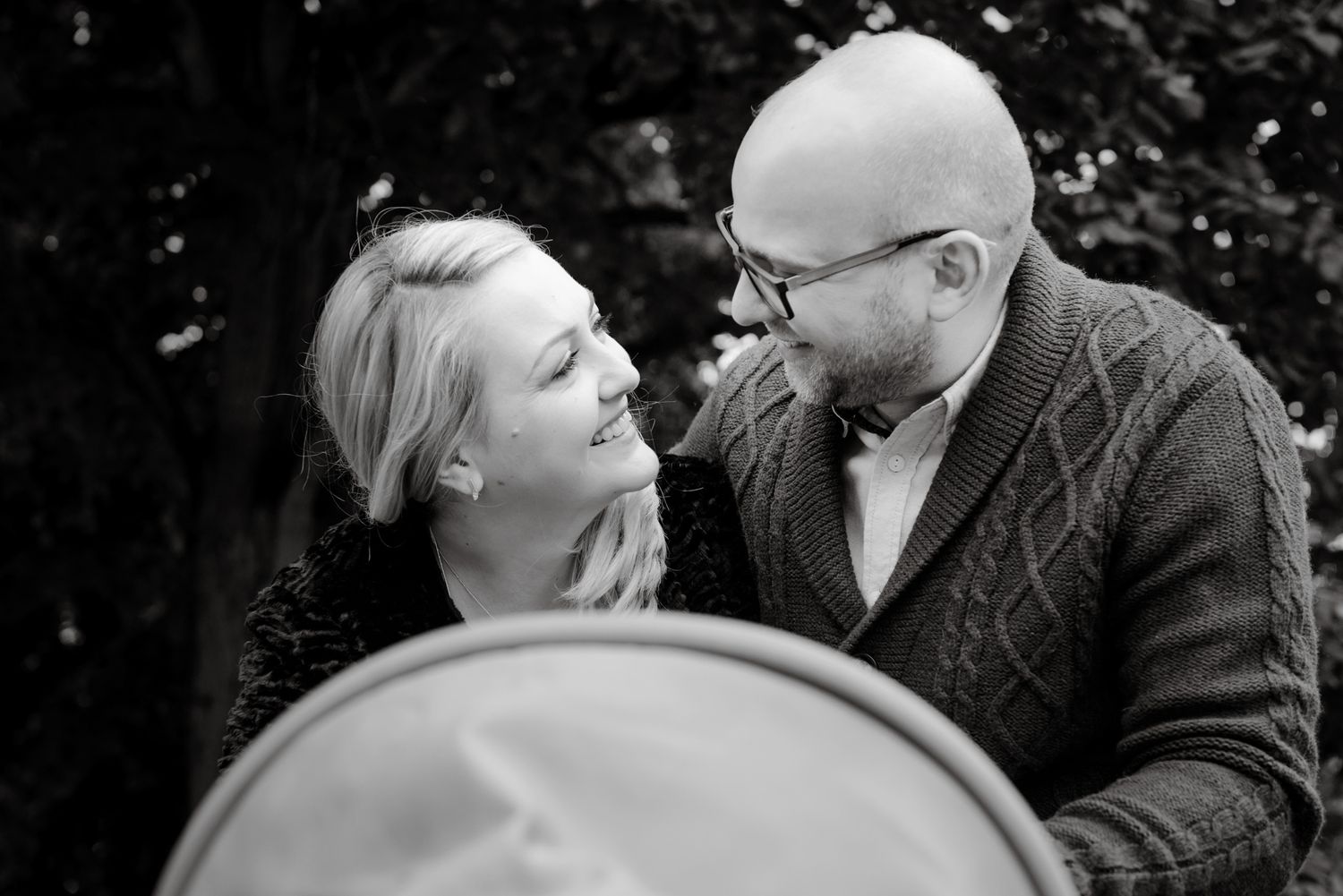 Black and white photo of a smiling couple gazing at each other outdoors, with a stroller in the foreground.