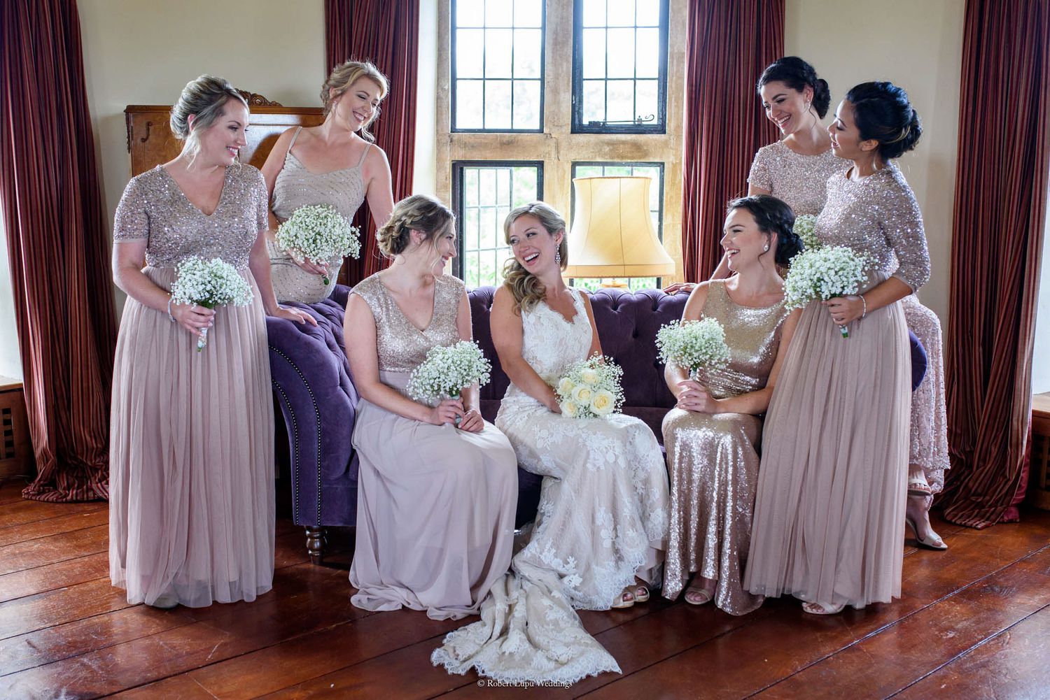 Bride in white lace gown poses with six bridesmaids in blush pink dresses holding white floral bouquets indoors.