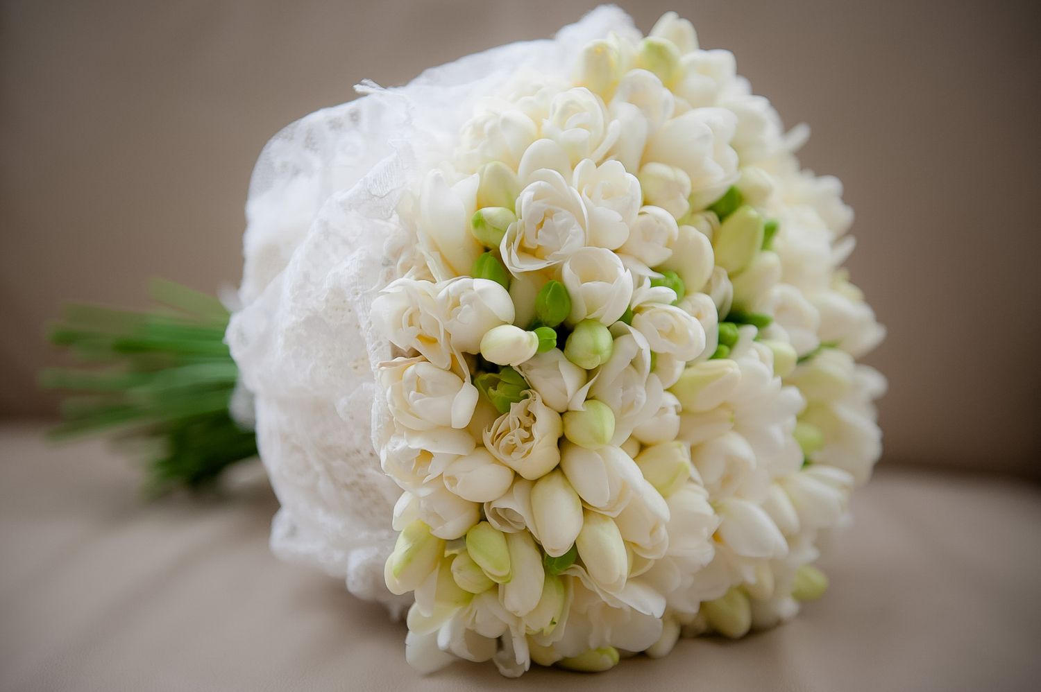 White bridal bouquet of tuberose flowers wrapped in lace ribbon, resting on a neutral background.