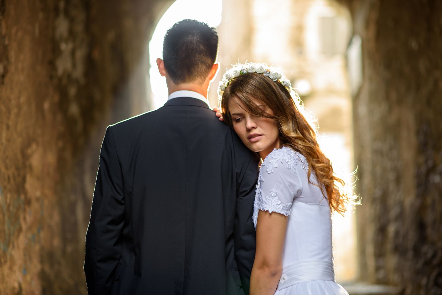 Bride in white dress resting her head on groom's shoulder in a romantic stone archway wedding photo.