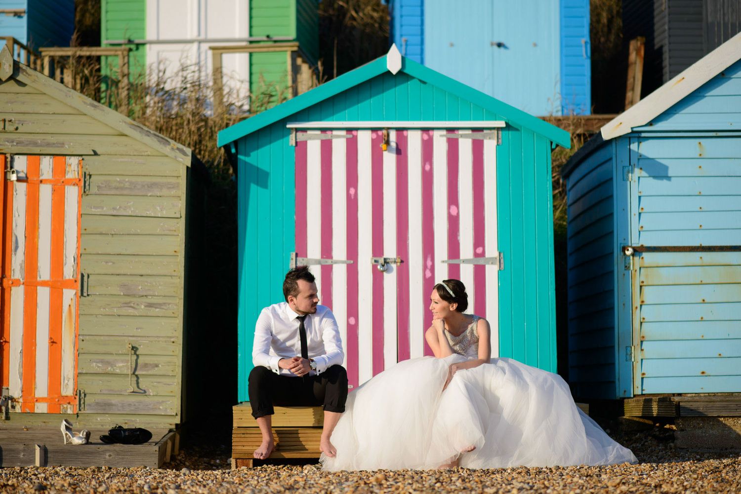 Bride and groom sitting together in front of colorful striped beach huts on a sunny wedding day.