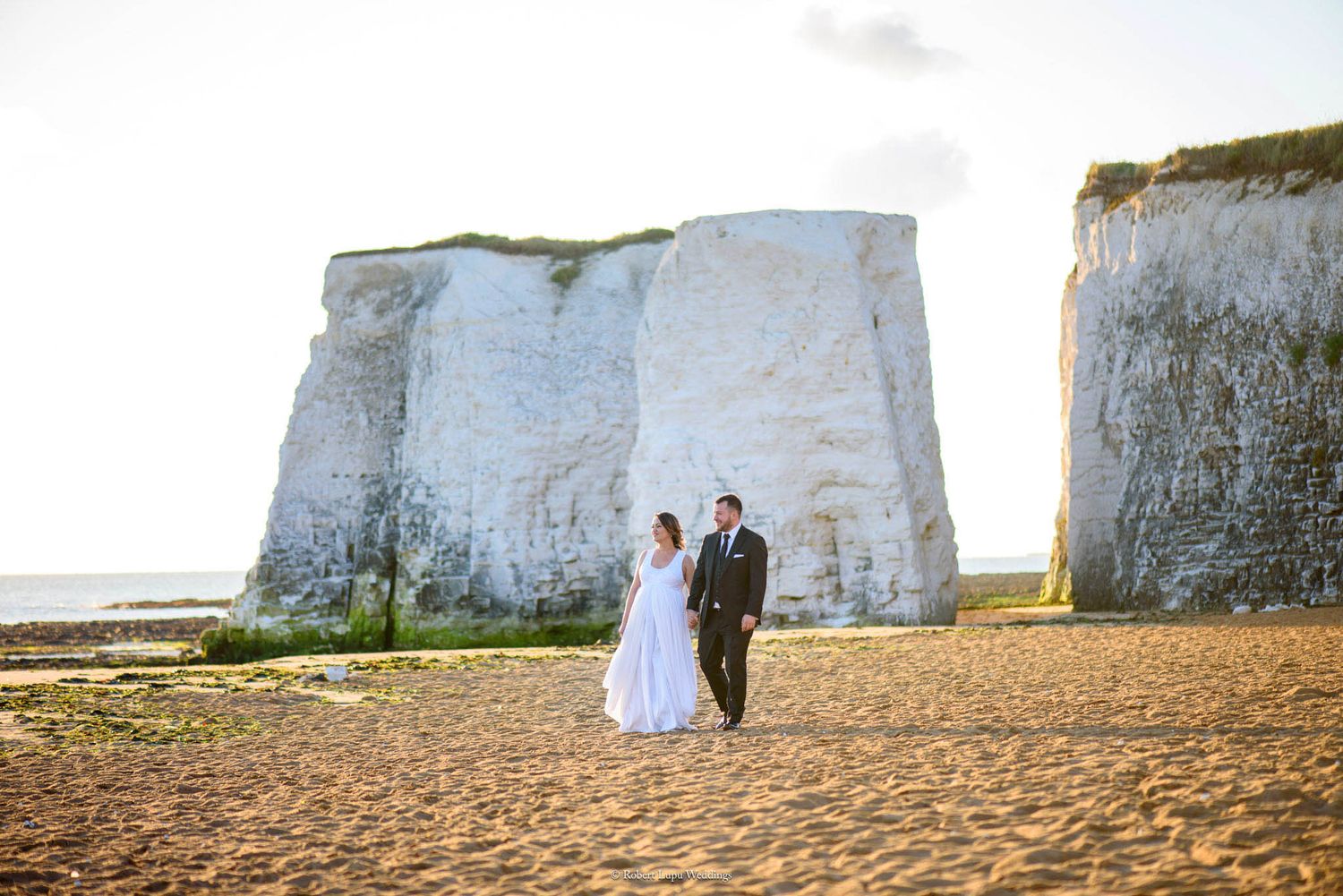 Bride and groom walking on sandy beach beside white chalk cliffs at golden hour.