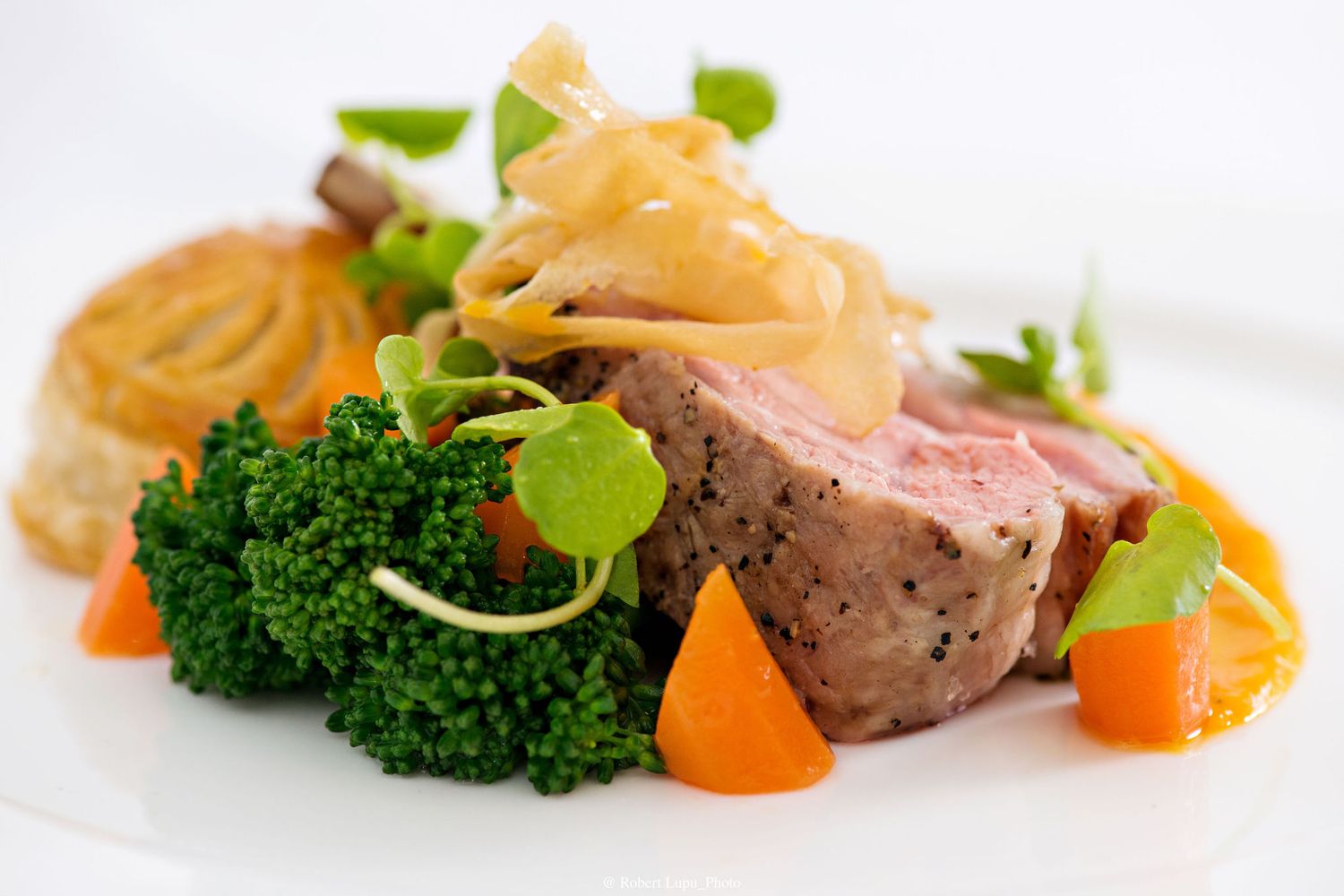 Elegant plated dish featuring sliced medium-rare meat, broccoli, carrots, caramelized onions, and a pastry on a white plate.