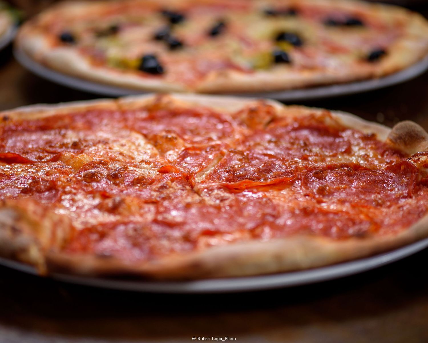 Classic pepperoni pizza with rich tomato sauce on a silver plate, with an olive pizza blurred in background.