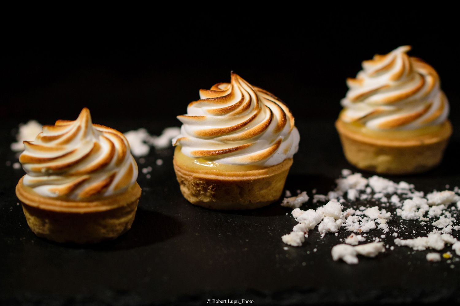 Three elegant lemon meringue tartlets with swirled toasted meringue tops on a dark slate surface.