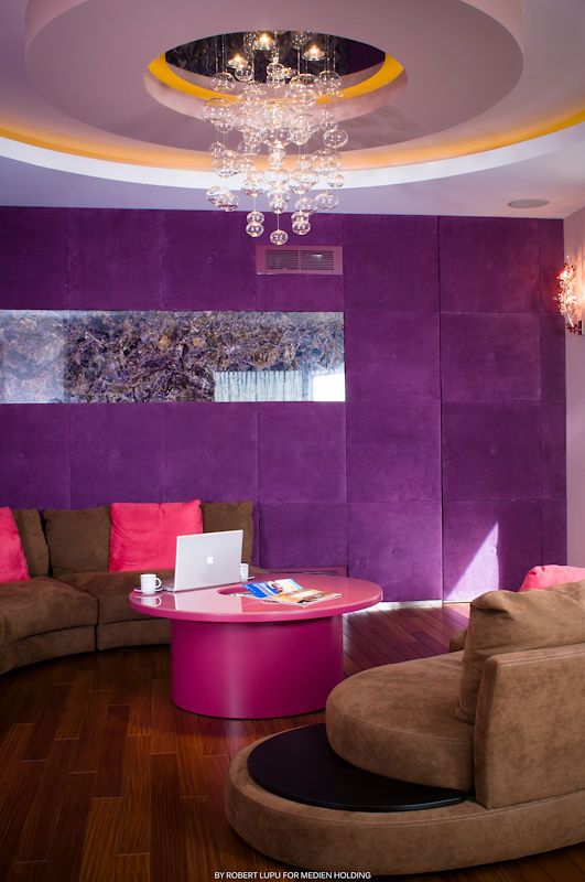 Modern lounge with purple velvet walls, pink coffee table, curved brown sofas, and a crystal bubble chandelier.