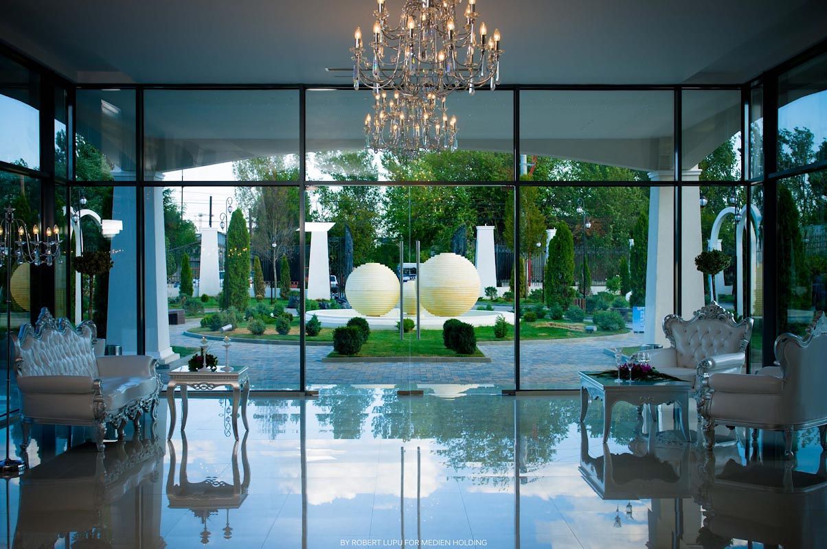 Elegant hotel lobby with glass walls, chandelier, white furniture, and illuminated spheres in a lush garden courtyard.