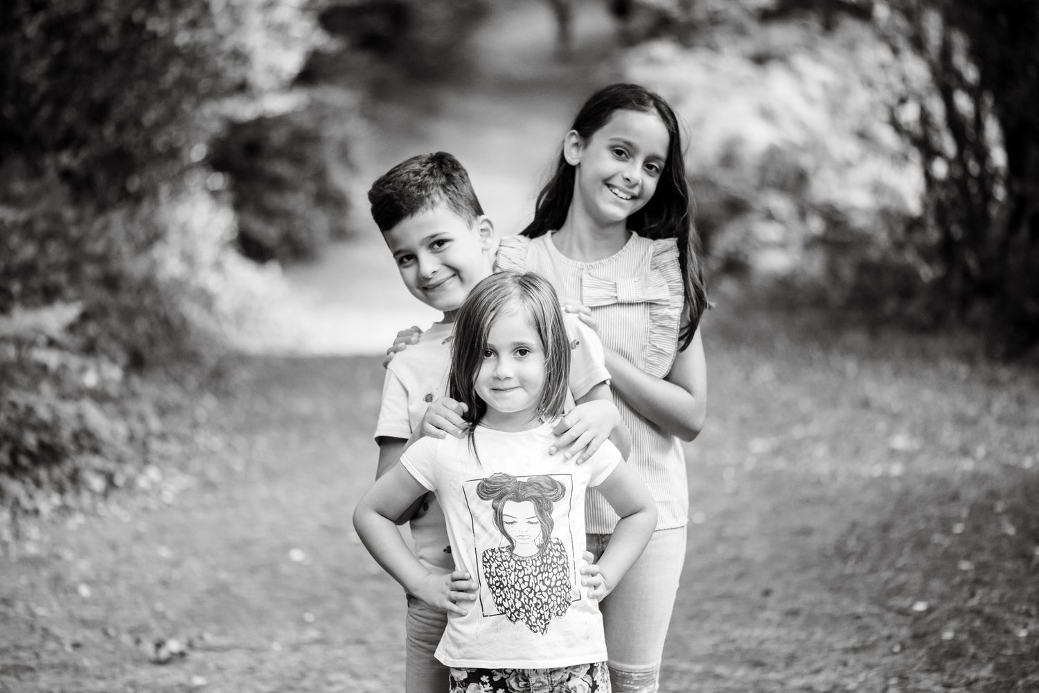 Black and white photo of three smiling children posing together on a park path surrounded by lush greenery.