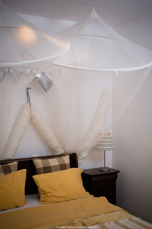 Romantic bedroom with yellow bedding, plaid pillows, white mosquito net canopy, and bedside lamp on dark wood nightstand.