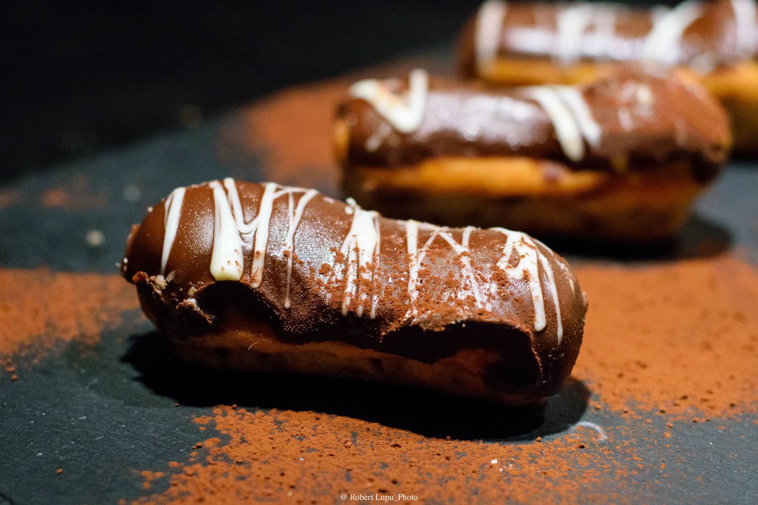 Chocolate eclairs topped with white drizzle on cocoa powder dusted surface.