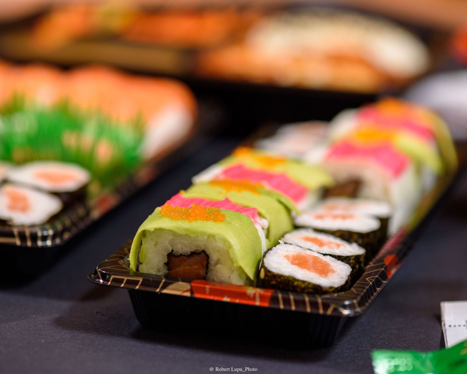 Colorful sushi rolls with avocado, tuna, and salmon displayed on black plastic trays at a Japanese food event.