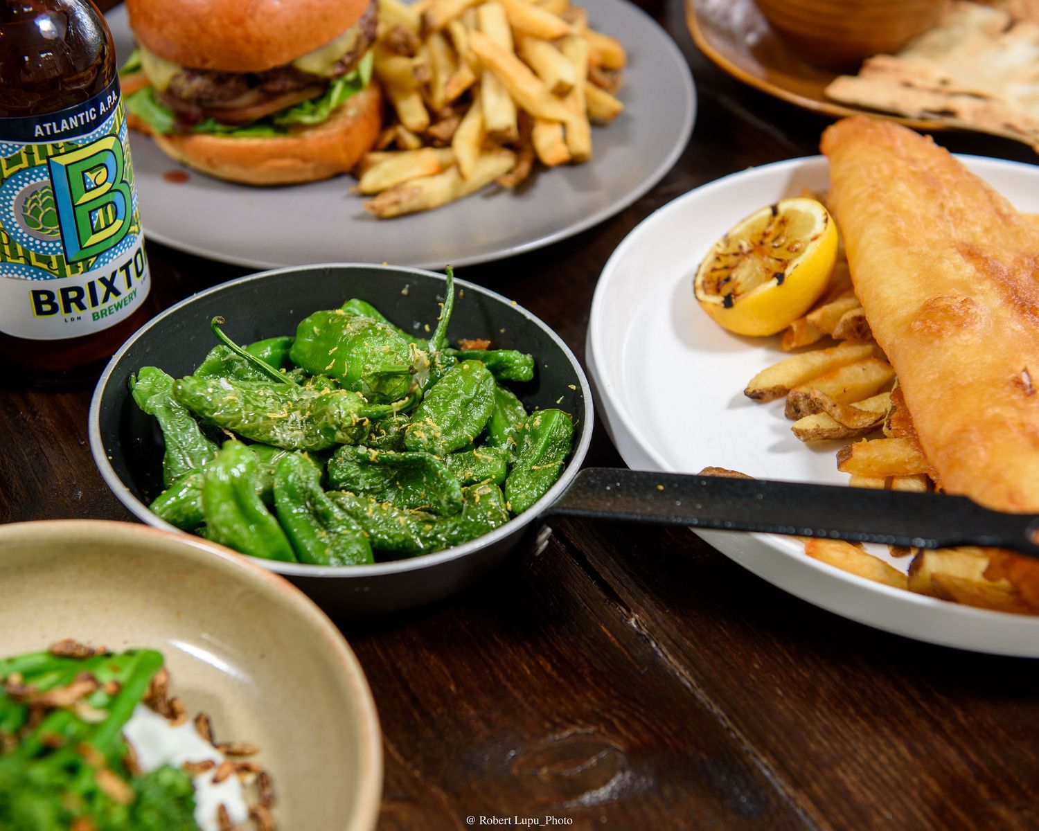 Spread of pub food including padron peppers, burger with fries, fish and chips, and Brixton beer bottle.