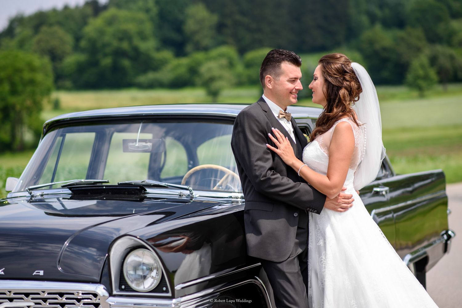 Bride and groom embracing beside a classic black vintage car outdoors on their wedding day.