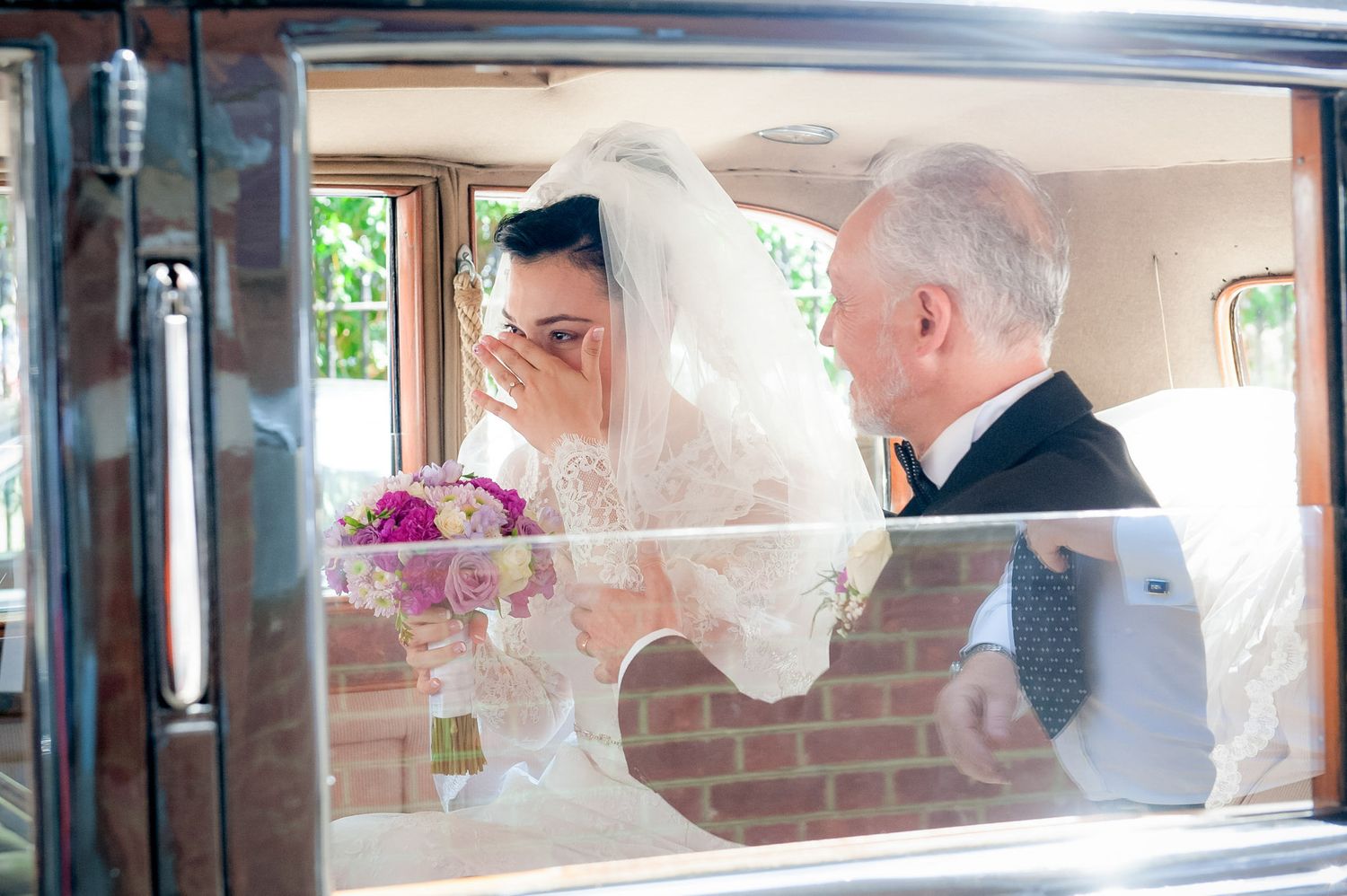 Emotional bride in white veil holding pink bouquet with older gentleman in suit, viewed through car window on wedding day.
