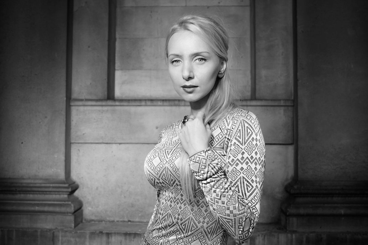 Elegant blonde woman in patterned dress posing against stone columns, black and white portrait.