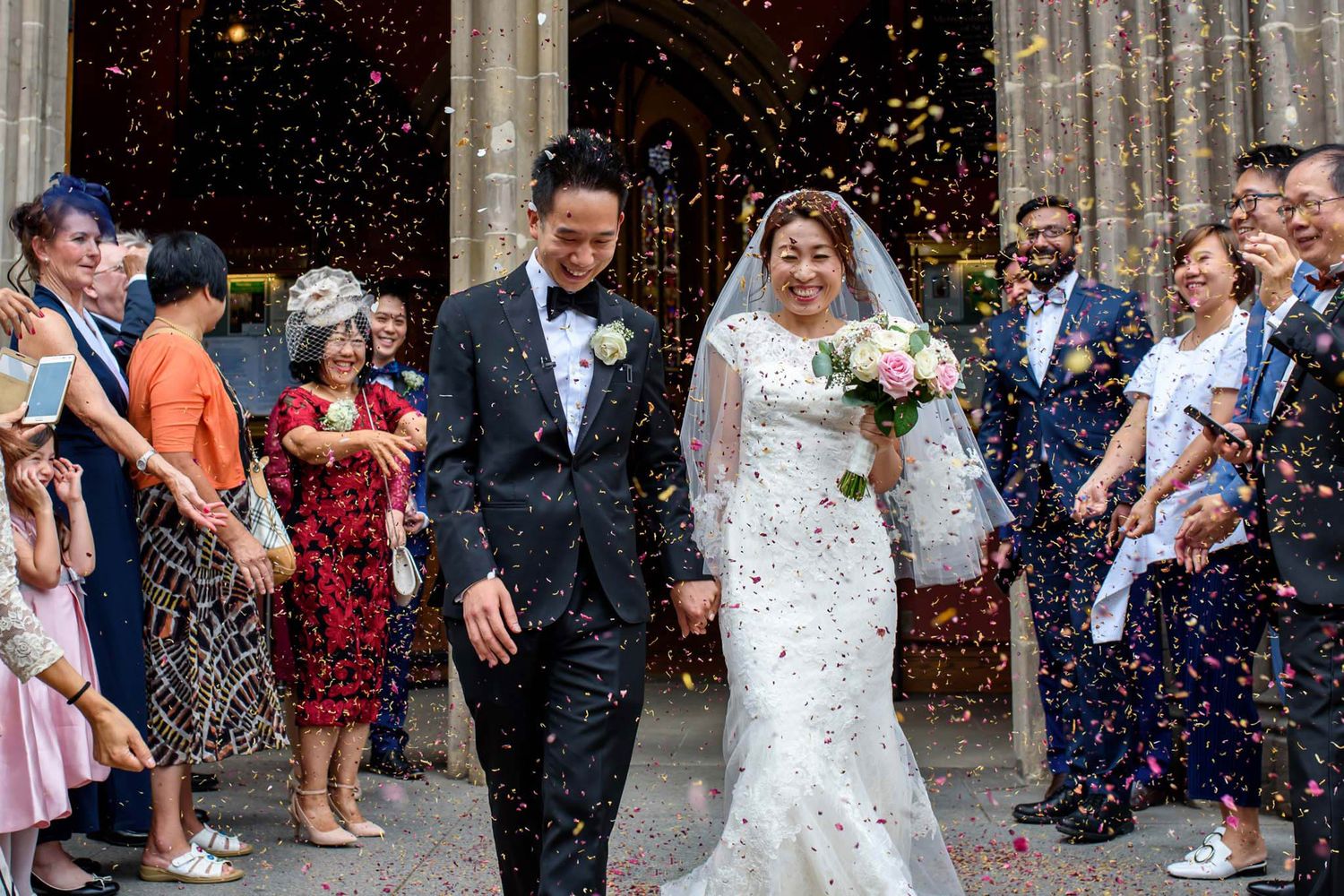 Newlywed Asian couple exits church as guests shower them with colorful confetti after their wedding ceremony.