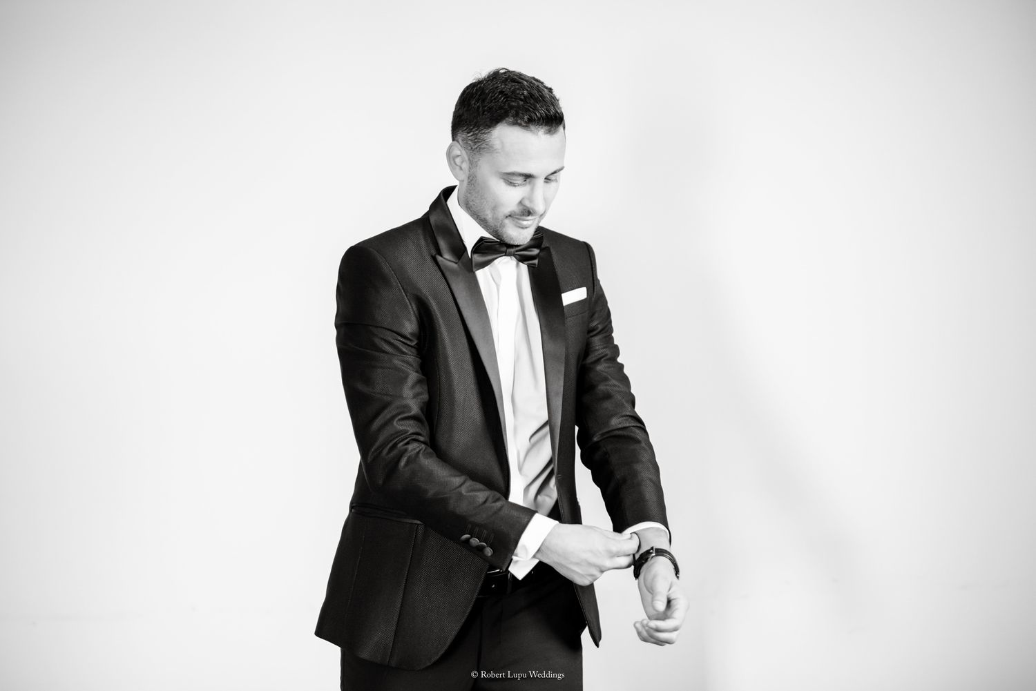 Black and white photo of a well-dressed man in a tuxedo adjusting his cufflinks against a white background.