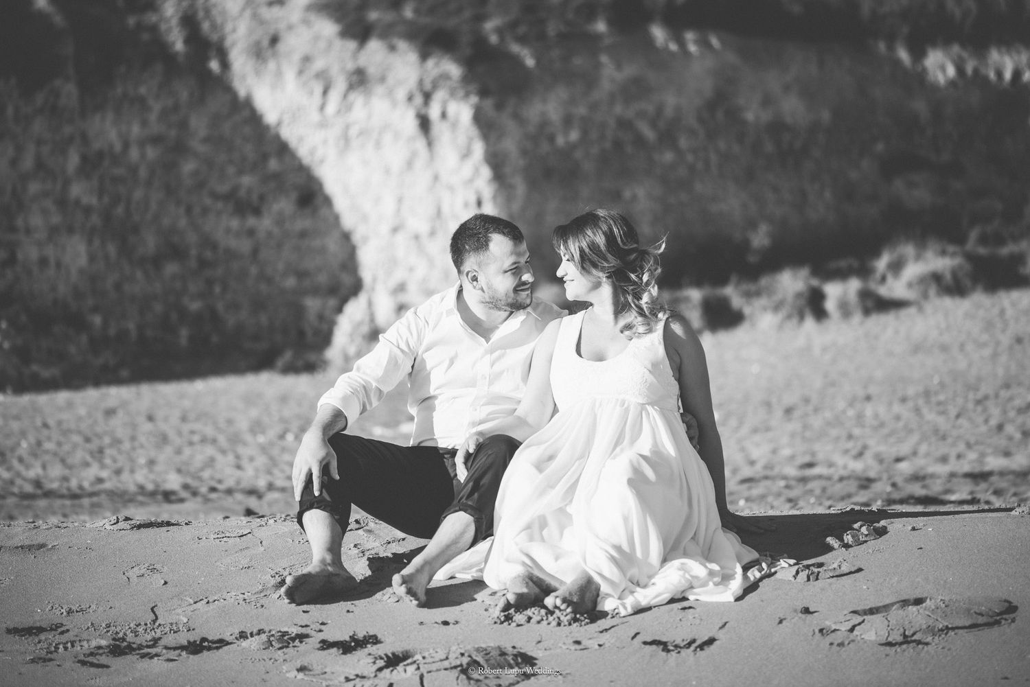 Black and white photo of a couple sitting together on a sandy beach, gazing at each other near rocky cliffs.