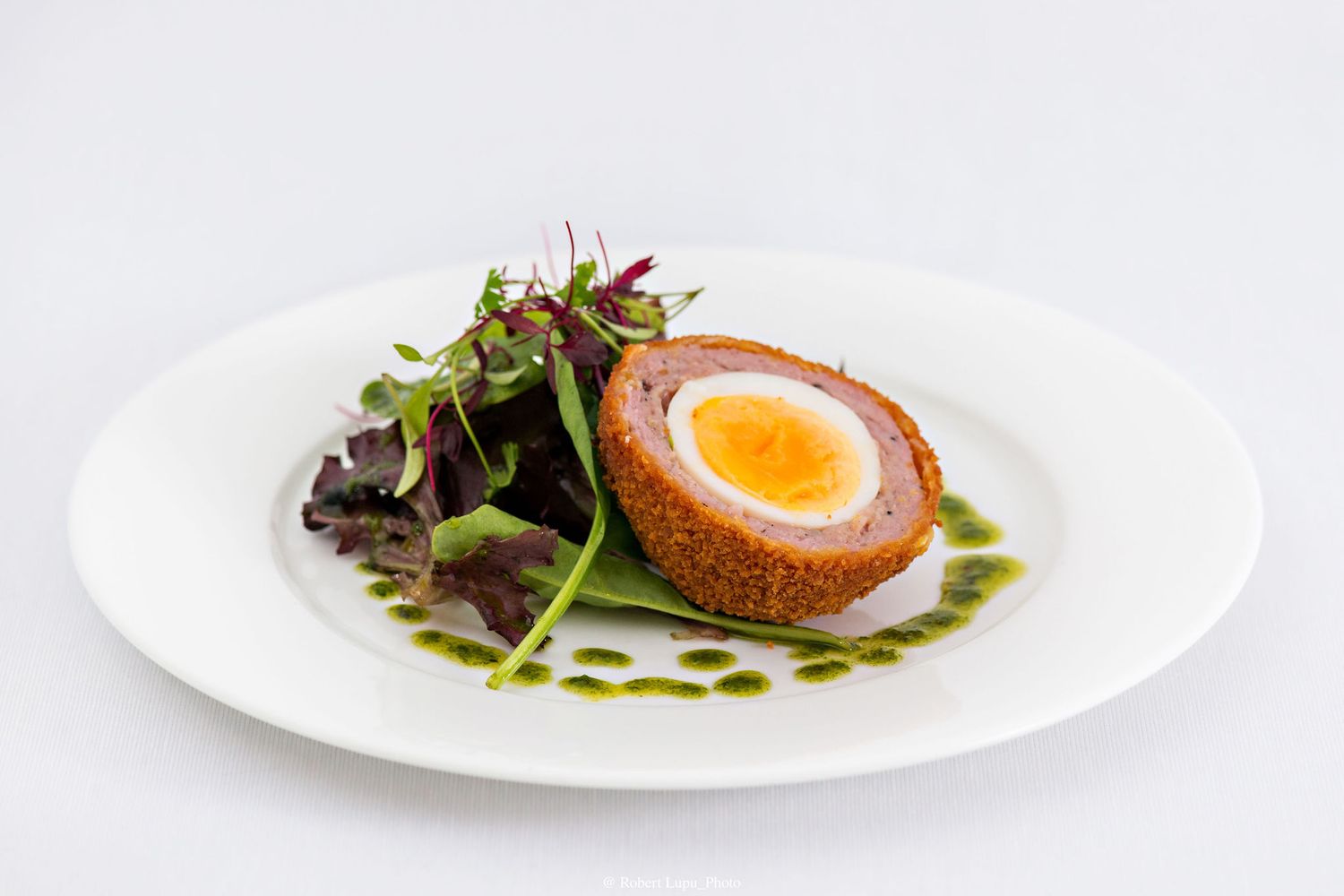 A classic Scotch egg with a soft yolk, breaded coating, mixed greens, and vibrant green herb sauce on a white plate.
