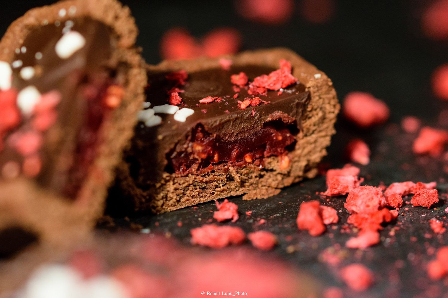 Decadent chocolate tart with raspberry filling, broken open to reveal layers, garnished with red candy pieces on dark slate.