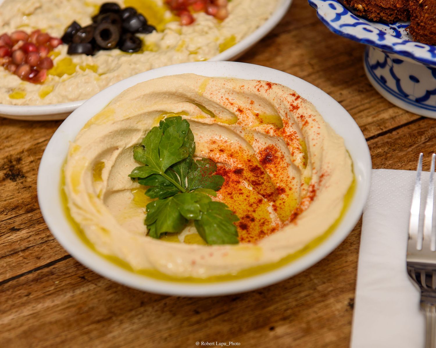 Creamy hummus garnished with fresh parsley, paprika, and olive oil served in a white bowl on a wooden table.