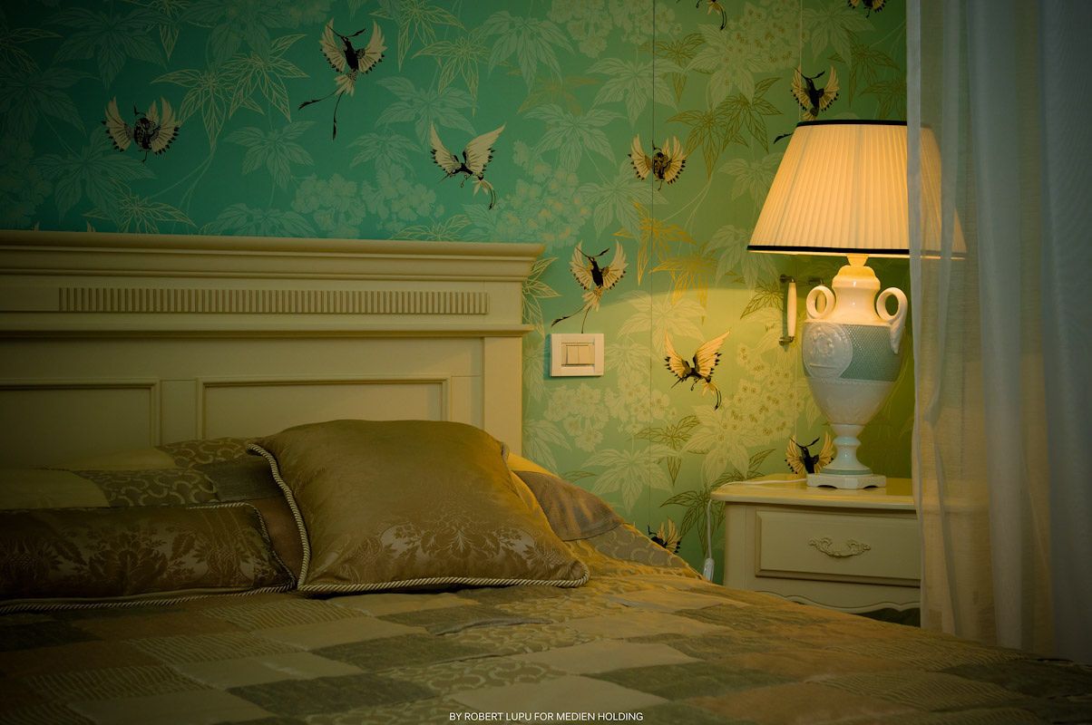 Romantic bedroom with teal butterfly wallpaper, white bed frame, and glowing vintage table lamp on nightstand.
