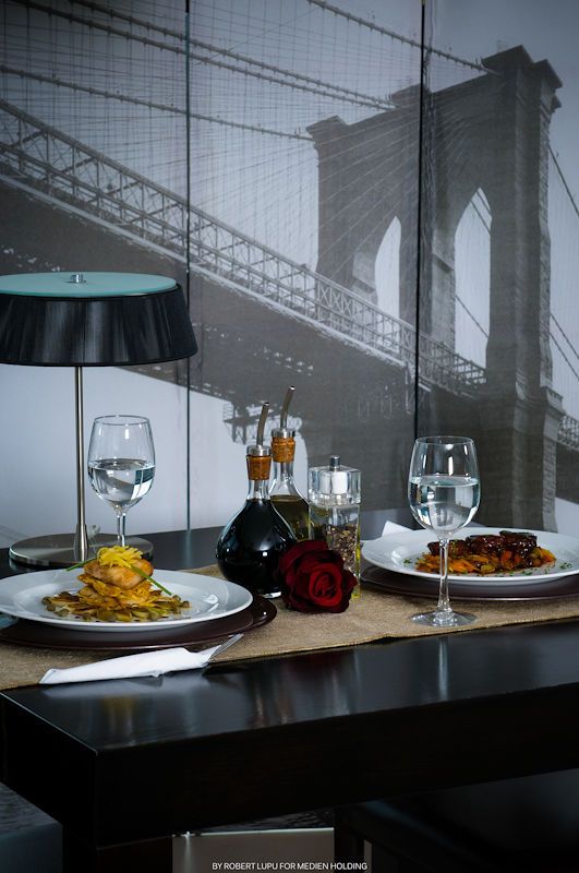 Elegant restaurant table set with pasta dishes, wine glasses, and a rose, with Brooklyn Bridge mural backdrop.