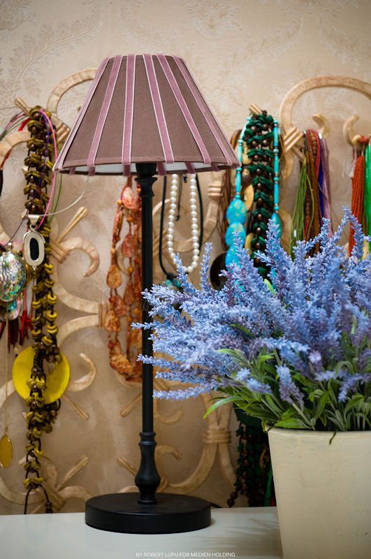Black table lamp with striped shade beside blue artificial lavender flowers and colorful necklaces on a decorative wall.
