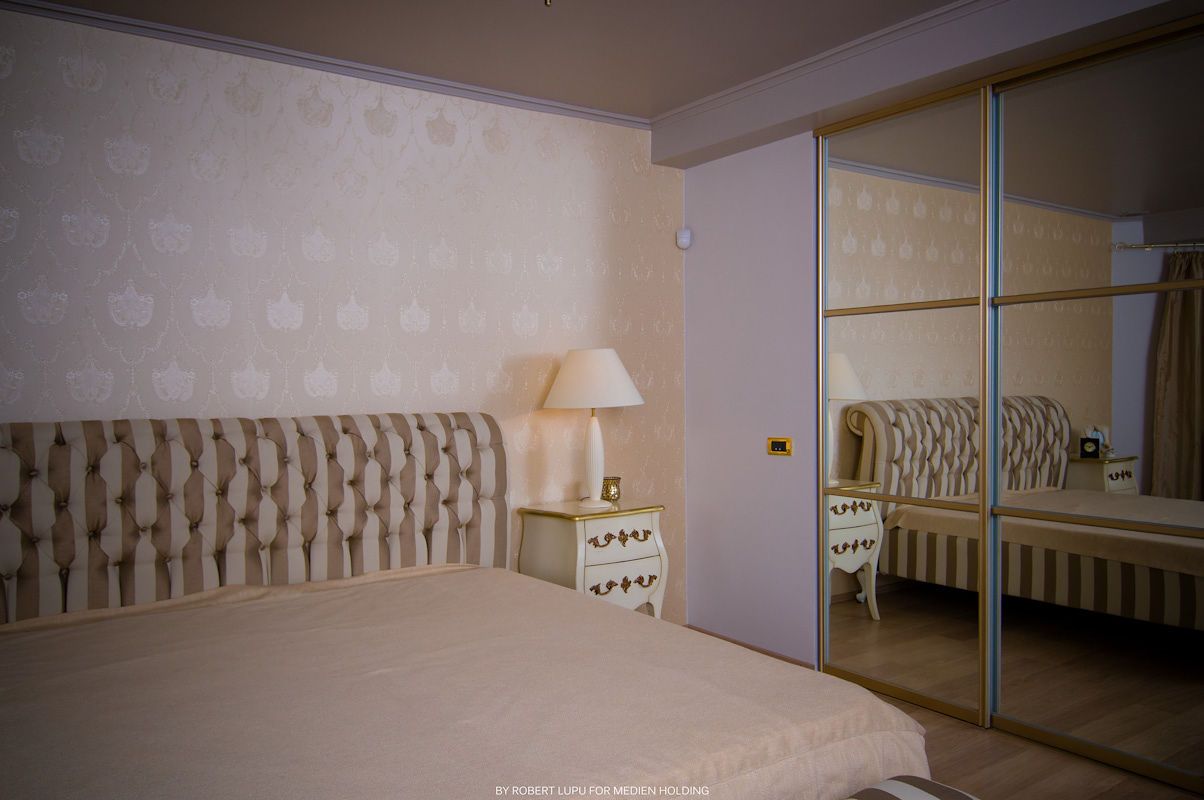Elegant bedroom with tufted headboard, mirrored wardrobe, bedside lamp, and floral wallpaper in soft neutral tones.