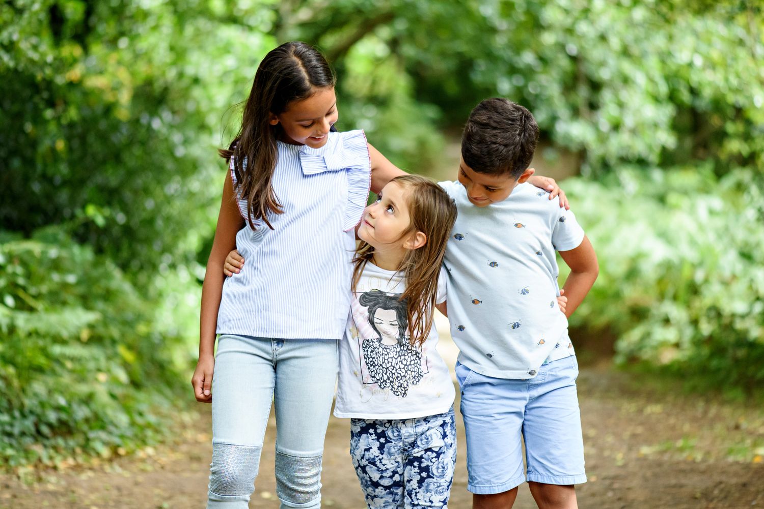 Three children hugging and smiling on a nature path surrounded by lush green trees.