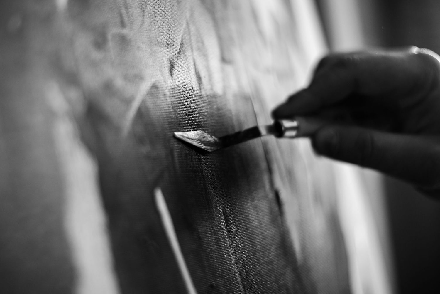 Black and white close-up of an artist's hand painting on canvas with a palette knife.