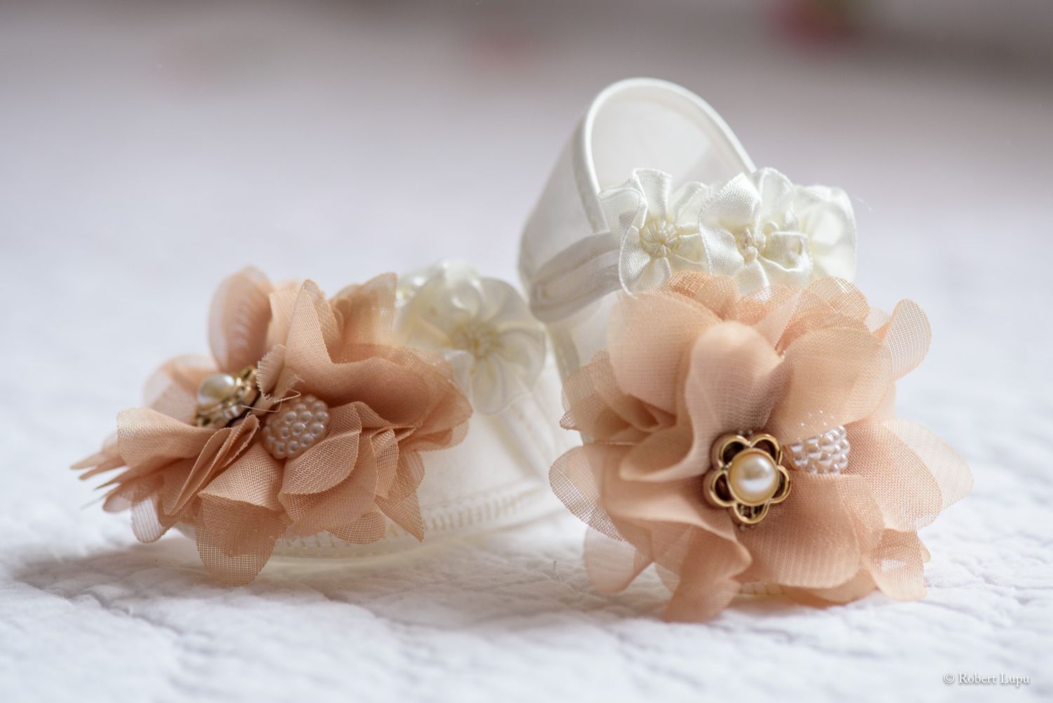 Delicate baby shoes adorned with chiffon flowers and pearl brooches in ivory and blush tones on lace.