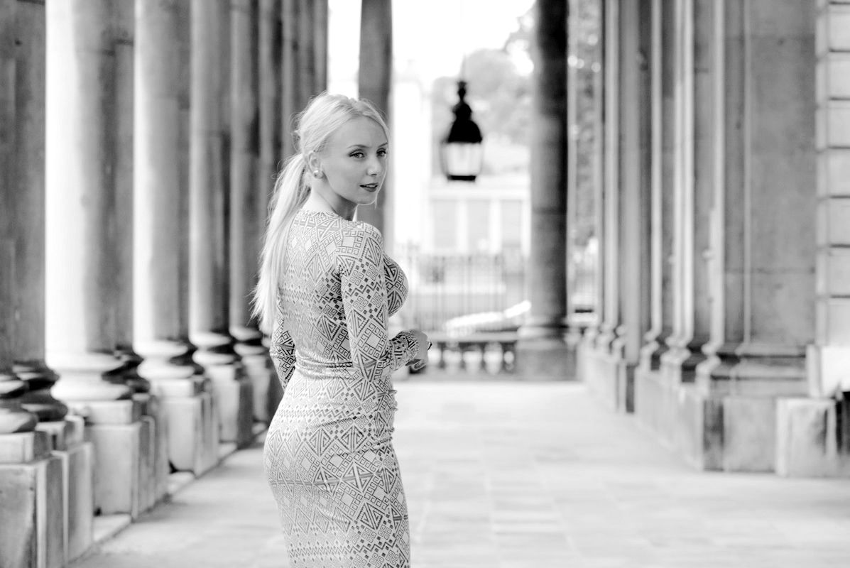 Blonde woman in elegant lace dress poses among classical columns in black and white architectural photo.