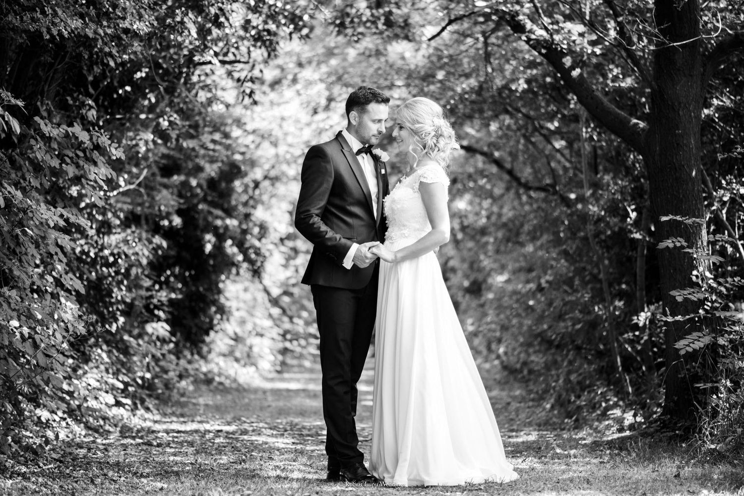 Bride and groom share a romantic moment on a tree-lined path in black and white wedding photo.