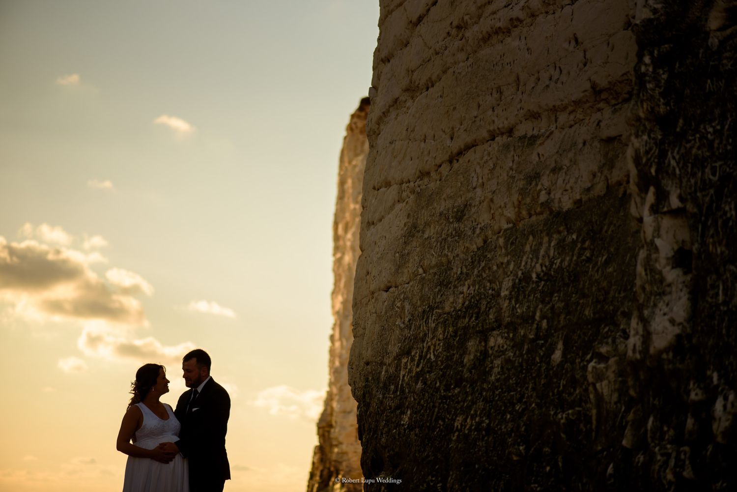 Couple silhouetted against golden sunset sky beside towering white chalk cliffs at romantic outdoor wedding photo shoot.