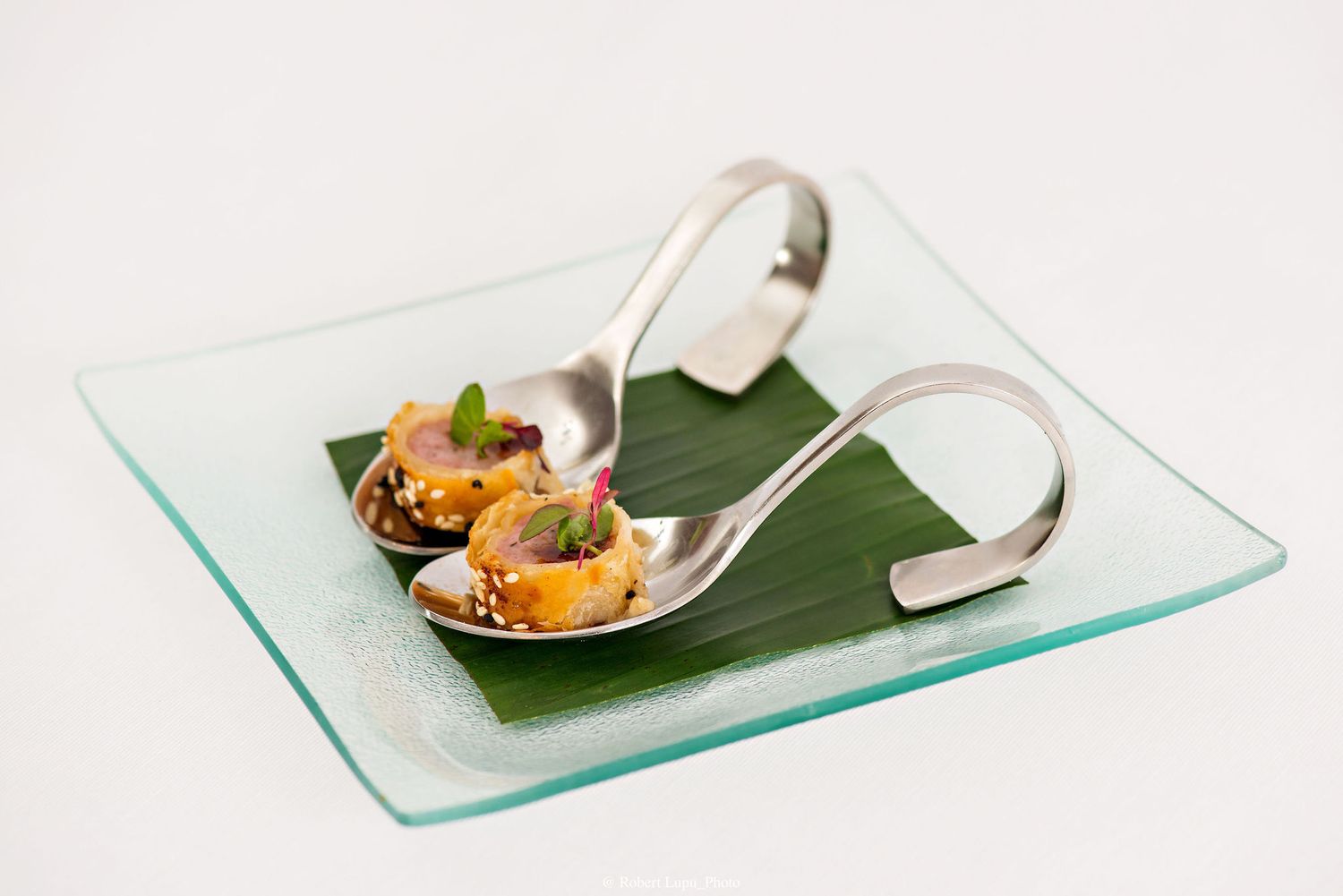 Two silver spoons with gourmet appetizers on a banana leaf atop a glass plate.